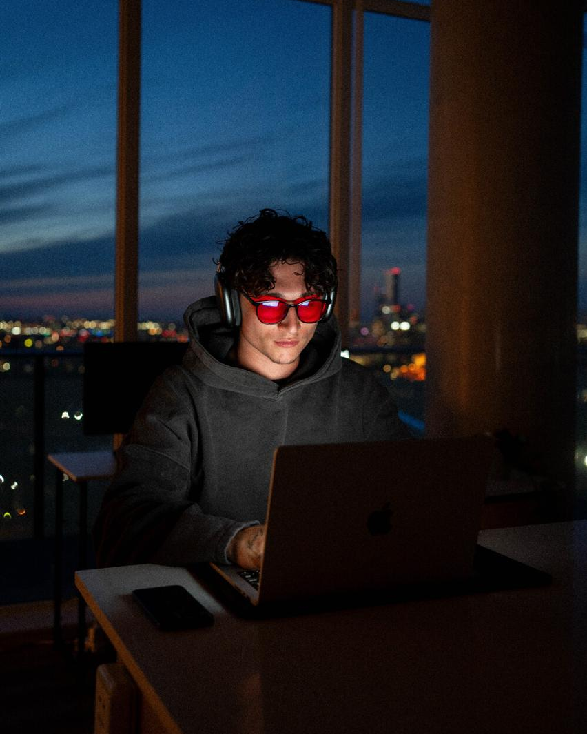 Person wearing red-tinted glasses and headphones while using a laptop by a window